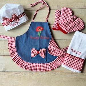 Puede incluir: Un delantal de cuadros rojos y blancos con un babero de mezclilla y una amapola roja bordada. El delantal tiene un bolsillo con un lazo de cuadros rojos y blancos. El texto "Poppy's Kitchen" está bordado en el delantal. El conjunto también incluye un gorro de chef de cuadros rojos y blancos, guantes para horno y una toalla de té de cuadros rojos y blancos con el nombre "Poppy" bordado.