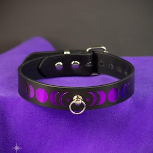 May include: Black leather collar with a purple moon phase design. The collar has a silver buckle and a metal ring. The design includes crescent moons and stars. The collar is displayed on purple fabric.