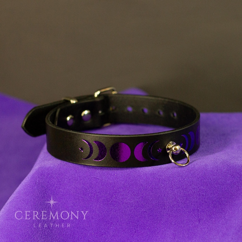 Pu&ograve; includere: Choker in pelle nera con fibbia argentata e un piccolo anello di metallo. Il choker presenta un design a fasi lunari viola. Il marchio "CEREMONY LEATHER" &egrave; stampato in basso nell'immagine.