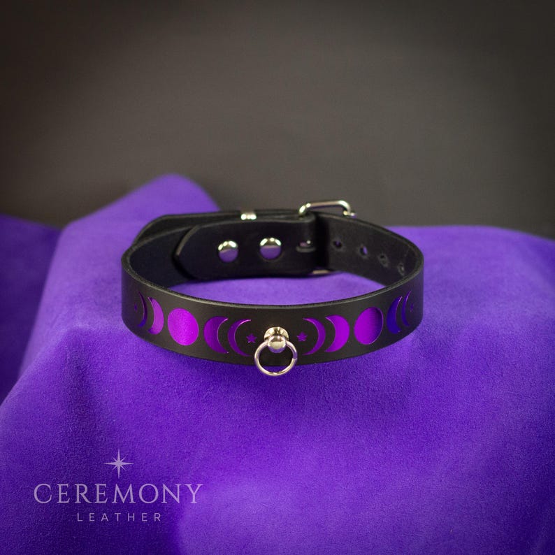 May include: Black leather collar with a purple moon phase design. The collar has a silver buckle and a metal ring. The design includes crescent moons and stars. The collar is displayed on purple fabric.