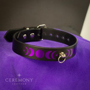 Pu&ograve; includere: Collare in pelle nera con fibbia argentata e anello a D. Il collare presenta un motivo ripetuto di lune crescenti e stelle viola. Il marchio "CEREMONY LEATHER" &egrave; stampato in bianco in basso.