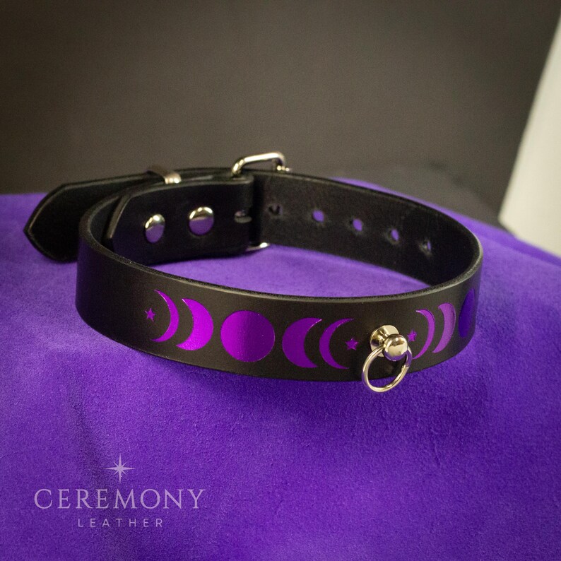 Pu&ograve; includere: Collare in pelle nera con fibbia argentata e anello a D. Il collare presenta un motivo ripetuto di lune crescenti e stelle viola. Il marchio "CEREMONY LEATHER" &egrave; stampato in bianco in basso.