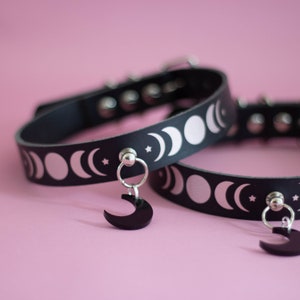 May include: Two black chokers with silver moon phase designs and a black crescent moon charm. The chokers have silver metal hardware and are set against a pink background. The chokers are approximately 1 inch wide.