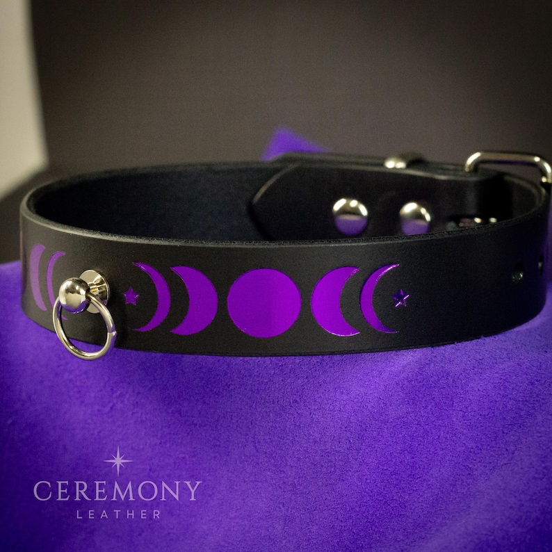 Pu&ograve; includere: Collare in pelle nera con design a fasi lunari viola. Presenta un anello e una fibbia in metallo argentato. Il testo "CEREMONY LEATHER" &egrave; stampato in basso. Larghezza di circa 4 cm.