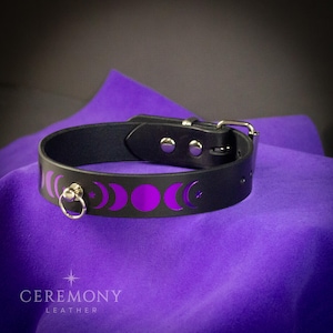 Pu&ograve; includere: Choker in pelle nera con fibbia e anello color argento. Il choker presenta un design a mezzaluna e stella viola. Il nome del marchio "CEREMONY LEATHER" &egrave; stampato in bianco in basso.
