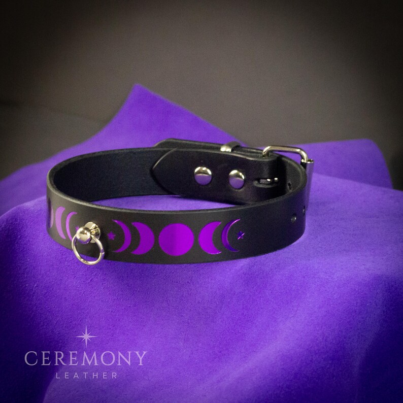 Pu&ograve; includere: Choker in pelle nera con fibbia e anello color argento. Il choker presenta un design a mezzaluna e stella viola. Il nome del marchio "CEREMONY LEATHER" &egrave; stampato in bianco in basso.
