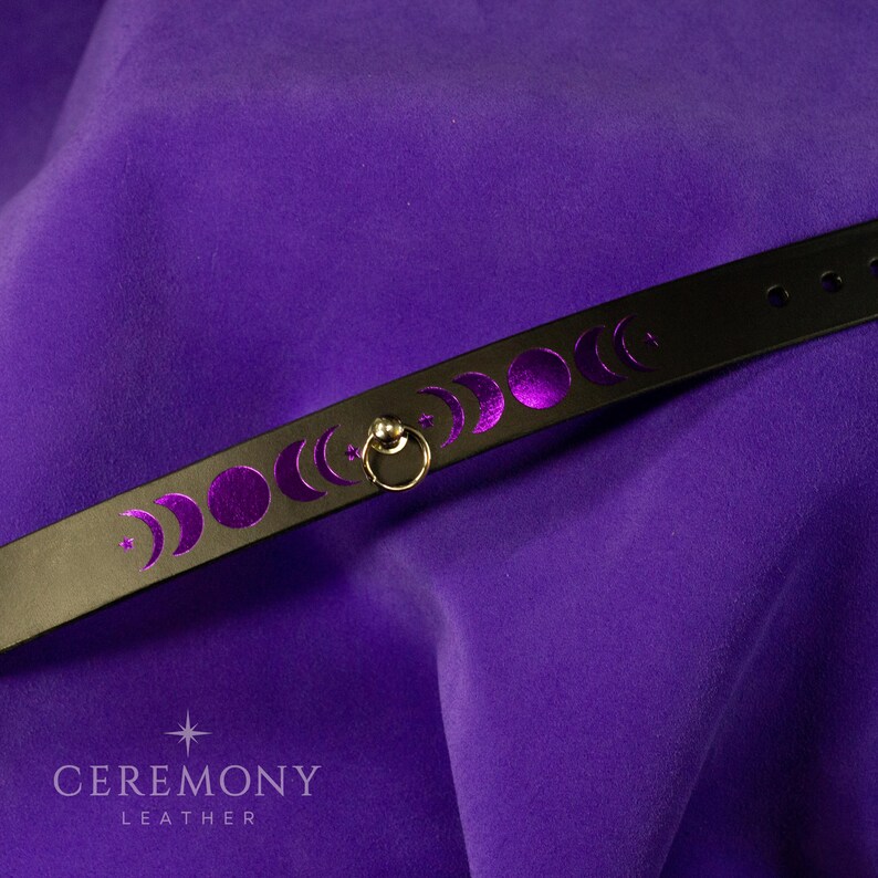 Pu&ograve; includere: Choker in pelle nera con fasi lunari viola. Un anello color oro &egrave; attaccato al choker. La parola "CEREMONY" &egrave; stampata in bianco, con "LEATHER" sotto. Sfondo viola.