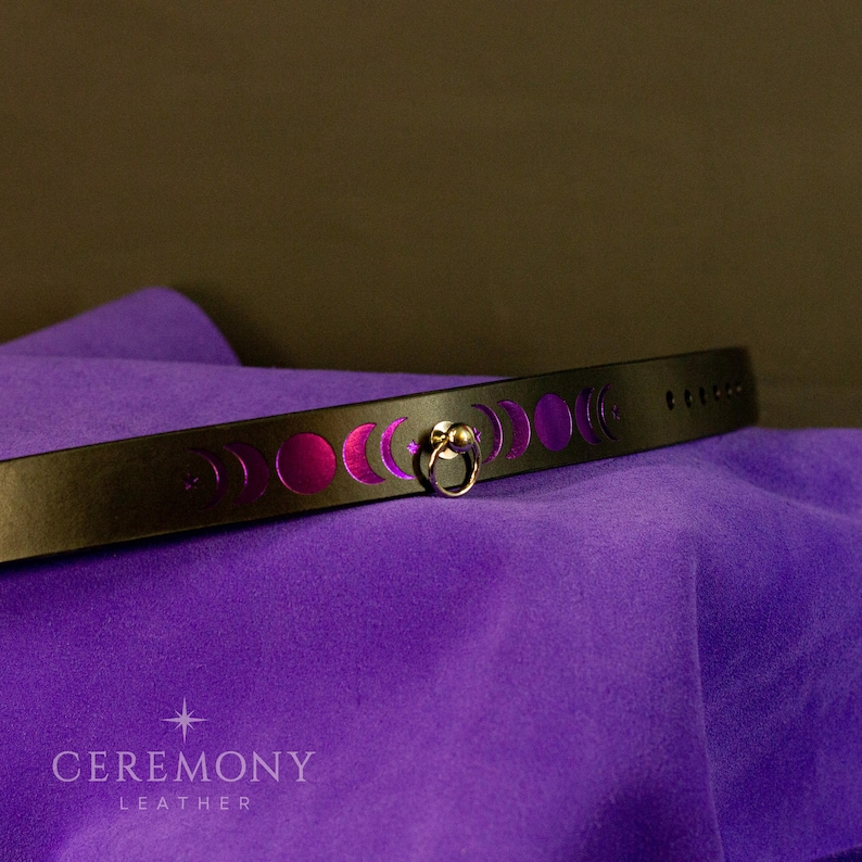 Pu&ograve; includere: Choker in pelle nera con design a fasi lunari e chiusura ad anello color oro. Il choker presenta ritagli di fasi lunari con un retro magenta. Il marchio "CEREMONY LEATHER" &egrave; stampato in basso.