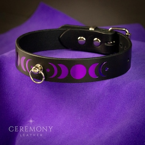 Pu&ograve; includere: Choker in pelle nera con fibbia color argento e anello a D. Il choker presenta un design a fasi lunari viola. Il testo "CEREMONY LEATHER" &egrave; in basso nell'immagine.