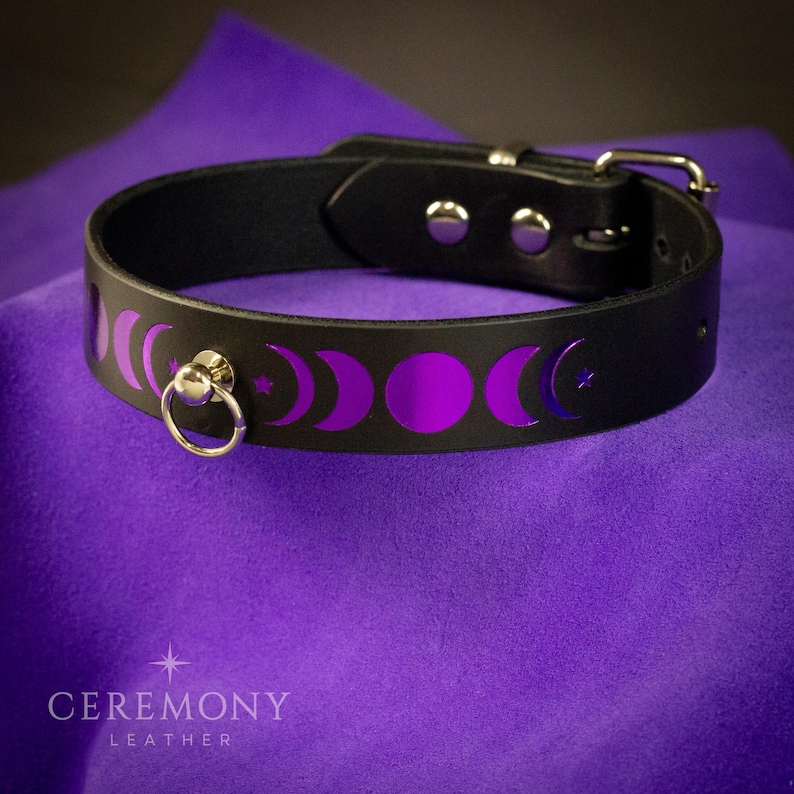 Pu&ograve; includere: Choker in pelle nera con fibbia color argento e anello a D. Il choker presenta un design a fasi lunari viola. Il testo "CEREMONY LEATHER" &egrave; in basso nell'immagine.