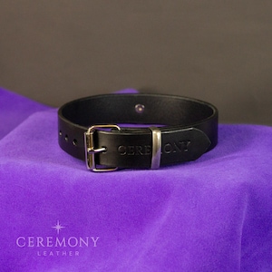 Pu&ograve; includere: Un braccialetto in pelle nera con fibbia argentata e la scritta "CEREMONY" in rilievo sul cinturino. Il braccialetto &egrave; esposto su una superficie di tessuto viola. Il marchio "CEREMONY LEATHER" &egrave; stampato in basso.