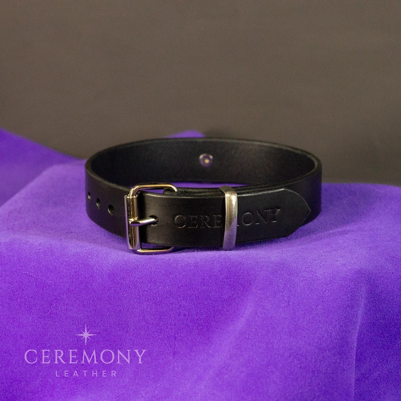 Pu&ograve; includere: Un braccialetto in pelle nera con fibbia argentata e la scritta "CEREMONY" in rilievo sul cinturino. Il braccialetto &egrave; esposto su una superficie di tessuto viola. Il marchio "CEREMONY LEATHER" &egrave; stampato in basso.