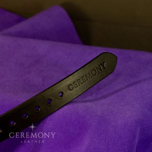 Pu&ograve; includere: Primo piano di una cintura in pelle nera con la scritta "CEREMONY" in rilievo sulla punta. La cintura presenta diversi fori distanziati e si staglia su uno sfondo di tessuto viola vibrante. &Egrave; visibile anche il marchio "CEREMONY LEATHER".