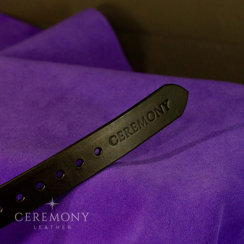 Pu&ograve; includere: Primo piano di una cintura in pelle nera con la scritta "CEREMONY" in rilievo sulla punta. La cintura presenta diversi fori distanziati e si staglia su uno sfondo di tessuto viola vibrante. &Egrave; visibile anche il marchio "CEREMONY LEATHER".