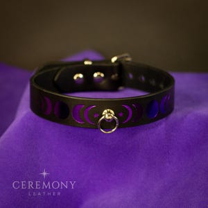 Pu&ograve; includere: Collare in pelle nera con ritagli a fasi lunari viola e un anello argentato. Il collare &egrave; fissato con una fibbia argentata. Il prodotto &egrave; presentato su una superficie viola. Il testo "CEREMONY LEATHER" &egrave; in basso.