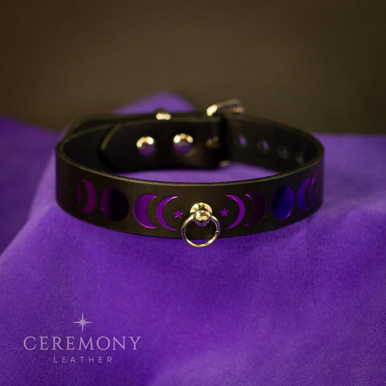 Pu&ograve; includere: Collare in pelle nera con ritagli a fasi lunari viola e un anello argentato. Il collare &egrave; fissato con una fibbia argentata. Il prodotto &egrave; presentato su una superficie viola. Il testo "CEREMONY LEATHER" &egrave; in basso.