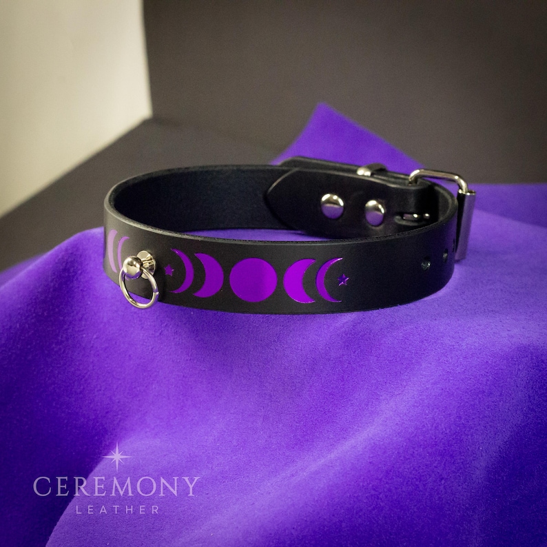 Pu&ograve; includere: Girocollo in pelle nera con design a fasi lunari viola. Presenta un anello e una chiusura con fibbia color argento. Il girocollo &egrave; esposto su tessuto viola. Il testo "CEREMONY LEATHER" &egrave; visibile in basso.