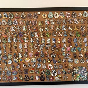 Può includere: Una bacheca incorniciata che mostra una vasta collezione di spille smaltate. Le spille presentano vari disegni colorati, tra cui personaggi dei cartoni animati e altre immagini fantasiose. Le spille sono attaccate alla bacheca con puntine.