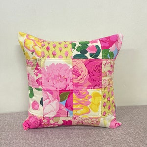 Op de afbeelding: Een vierkant kussen met een patchwork-ontwerp met roze, gele en groene bloemenpatronen. Het kussen is gemaakt van stof en heeft een witte achtergrond.
