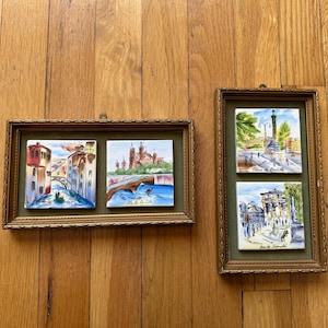 May include: Three framed ceramic tiles with watercolor paintings of different scenes in Paris, France. The tiles are in gold frames and are hung on a wooden wall. The tiles depict the Place de la Bastille, Ile de la Cité, and a street scene.