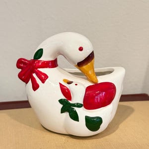 May include: White ceramic goose planter with a red bow and green leaves. The goose is holding a smaller goose in its beak. The planter is perfect for small plants or flowers.