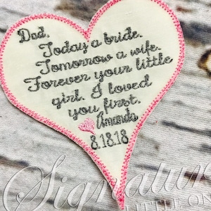 May include: A white heart-shaped fabric patch with pink stitching around the edges. The patch has a message embroidered in black thread that reads: "Dad. Today a bride. Tomorrow a wife. Forever your little girl. I loved you first. Amanda 8.18.18"