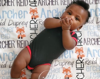 May include: A baby boy wearing a black bodysuit with red trim is lying on a white blanket with a fox print. The blanket has the names "Archer Reid" and "Reid Duprey" printed on it in black and gray.