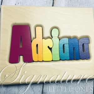 May include: A wooden puzzle with the name "Adriana" spelled out in rainbow colors. The letters are raised and can be removed from the board. The board is made of light wood and has the words "Signature Little Ones" printed on the bottom.
