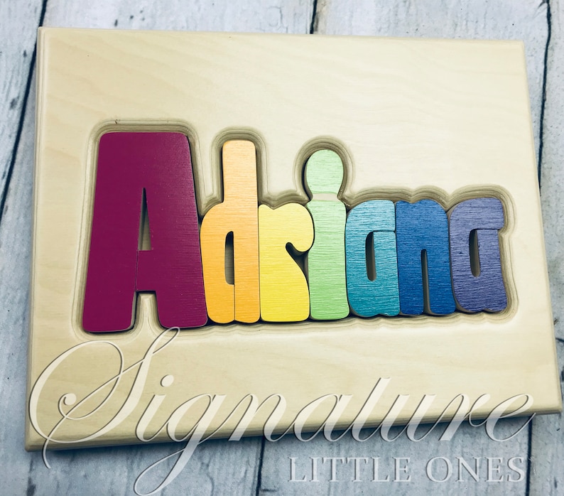 May include: A wooden puzzle with the name "Adriana" spelled out in rainbow colors. The letters are raised and can be removed from the board. The board is made of light wood and has the words "Signature Little Ones" printed on the bottom.