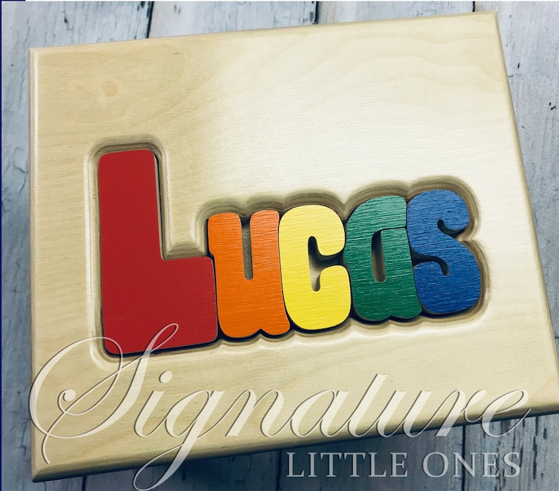 May include: A wooden puzzle with the name "Lucas" spelled out in colorful letters. The letters are red, orange, yellow, green, and blue. The puzzle is on a light wood background with the words "Signature Little Ones" printed below the puzzle.