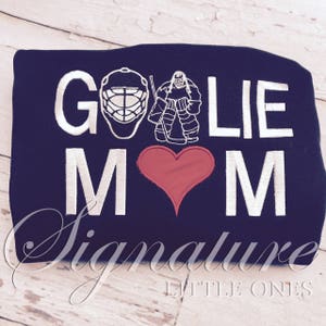 Pu&ograve; includere: Una felpa blu navy con ricamo bianco che recita "Goalie Mom" con un cuore rosso al centro. Il ricamo include anche un portiere di hockey con un bastone e una maschera da hockey.