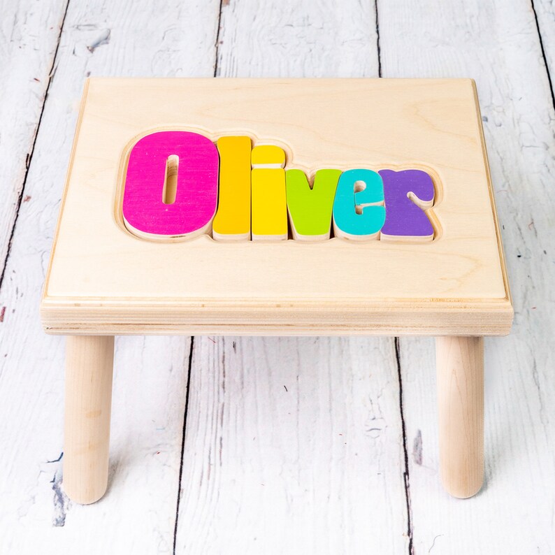 Op de afbeelding: Een houten opstapje met de naam "Oliver" gespeld in kleurrijke letters op de bovenkant. De letters zijn roze, geel, groen, blauw en paars.