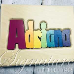 Op de afbeelding: Een houten puzzel met de naam "Adriana" gespeld in regenboogkleuren. De letters zijn verhoogd en kunnen van het bord worden verwijderd. Het bord is gemaakt van licht hout en heeft de woorden "Signature Little Ones" onderaan gedrukt.
