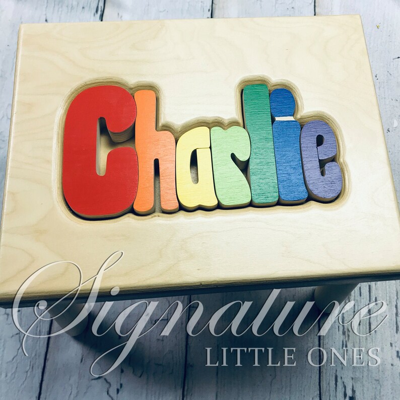 Op de afbeelding: Een houten opstapje met de naam "Charlie" gespeld in puzzelstukjes in regenboogkleuren. De kruk is gemaakt door Signature Little Ones.