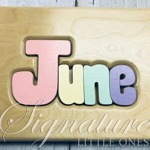 May include: A wooden puzzle with the name "June" spelled out in colorful letters. The letters are pink, yellow, green, and purple. The puzzle is made by Signature Little Ones.