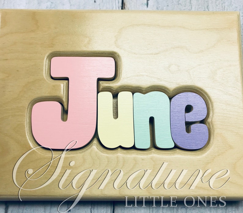 May include: A wooden puzzle with the name "June" spelled out in colorful letters. The letters are pink, yellow, green, and purple. The puzzle is made by Signature Little Ones.