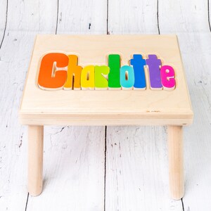 Op de afbeelding: Een houten opstapje met de naam "Charlotte" gespeld in kleurrijke letters. De letters zijn in een regenboogpatroon, met elke letter een andere kleur.