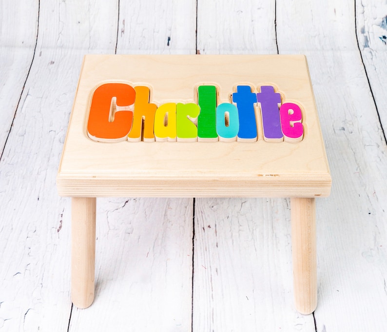 Op de afbeelding: Een houten opstapje met de naam "Charlotte" gespeld in kleurrijke letters. De letters zijn in een regenboogpatroon, met elke letter een andere kleur.
