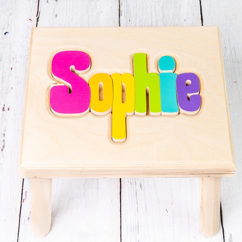 May include: A wooden step stool with the name "Sophie" spelled out in colorful letters. The letters are pink, yellow, green, blue, and purple.