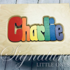 Op de afbeelding: Een houten puzzel met de naam "Charlie" gespeld in regenboogkleuren. De puzzel is gemaakt van afzonderlijke stukken die in elkaar passen. De puzzel ligt op een houten basis met de woorden "Signature Little Ones" gegraveerd aan de onderkant.