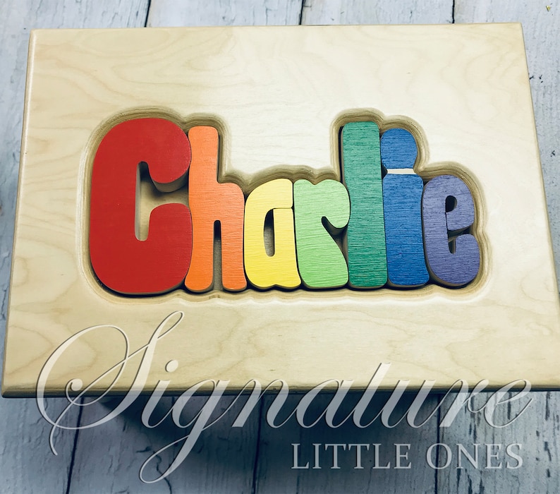 Op de afbeelding: Een houten puzzel met de naam "Charlie" gespeld in regenboogkleuren. De puzzel is gemaakt van afzonderlijke stukken die in elkaar passen. De puzzel ligt op een houten basis met de woorden "Signature Little Ones" gegraveerd aan de onderkant.