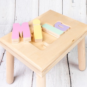 May include: A wooden step stool with four legs and a top surface that has the letters "MILA" cut out. The letters are painted in pink, yellow, and green.