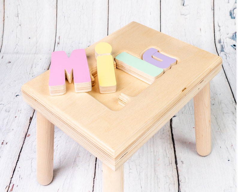 May include: A wooden step stool with four legs and a top surface that has the letters "MILA" cut out. The letters are painted in pink, yellow, and green.