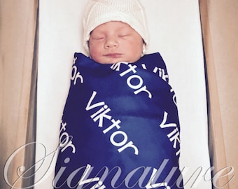 May include: A newborn baby wearing a white knitted hat and a blue swaddle blanket with the name "Viktor" printed on it.