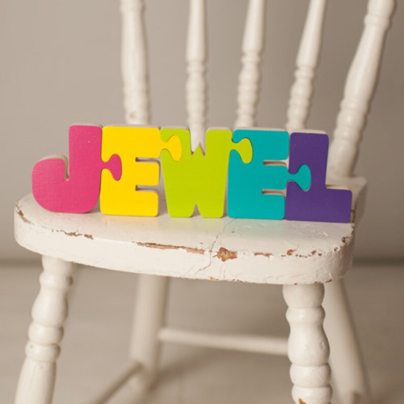 Op de afbeelding: Kleurrijke houten puzzelstukjes die de naam "JEWEL" spellen op een witte, verweerde houten stoel.