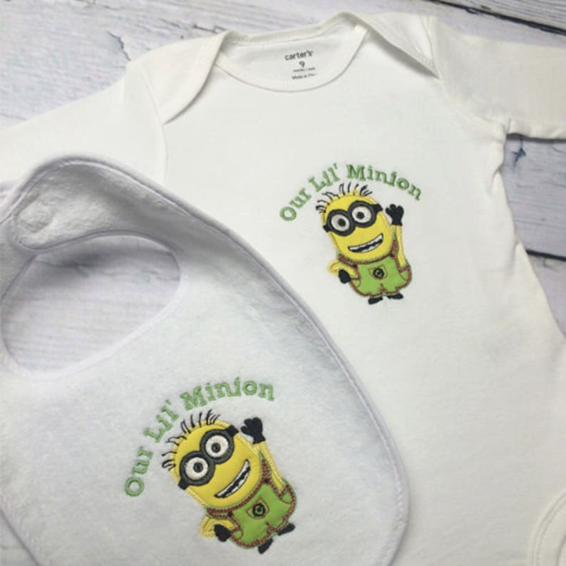 May include: White baby onesie and bib with an embroidered yellow minion character. The text "Our Lil' Minion" is embroidered in green on both items.