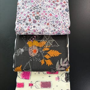 May include: Three folded fabric items with unique patterns. The top item features a floral design with skulls. The middle item has a black background with a floral and spiderweb design. The bottom item displays a pattern of potion bottles.
