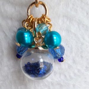 May include: A gold necklace with a charm featuring a clear glass orb filled with blue glitter. The charm is surrounded by blue and turquoise beads.