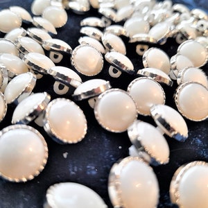 May include: A large group of white pearl buttons with silver trim. The buttons are scattered on a dark blue surface.