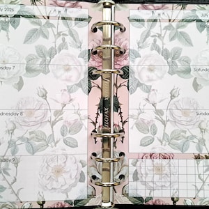 May include: A planner insert for July 2026, featuring a floral rose design in pink, white, and green. The weekly layout includes dates and days of the week, with a grid for notes. The planner is held together by a silver binder.